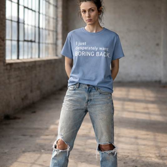 A womain standing in an empty industrial loft looking directly at the camera with her hands behind her back. She is wearing a  washed denim colored graphic t-shirt with the words "I just desperately want BORING BACK." printed in bold white sans-serif text across the chest and worn blue jeans with torn knees. 