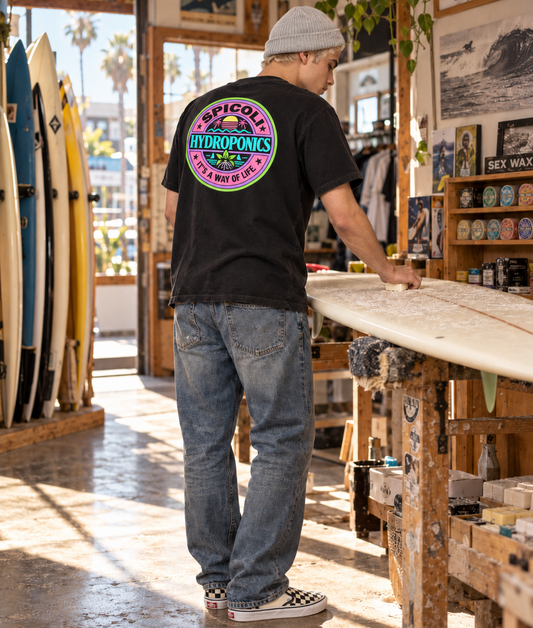 A young man in a gray knit beanie stands inside a SoCal surf shop, seen mostly from behind as he applies wax to a surfboard. He wears a black Spicoli Hydroponics T-shirt with a large colorful circular back graphic, worn blue jeans, and black-and-white checkerboard slip-on shoes. Surfboards, wax displays, posters, and warm natural light fill the shop around him.