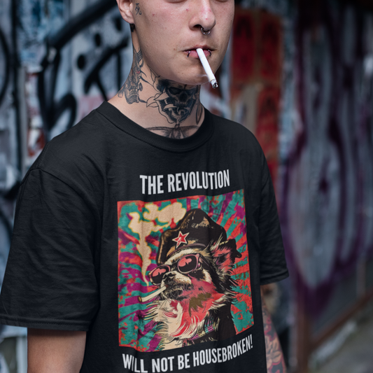 A man standing against a graffiti background wearing a black colored short-sleeve crewneck t-shirt featuring a vibrant, pop-art style square graphic on the chest. The illustration shows a long-haired Chihuahua stylized as a revolutionary, wearing a black hat with a red star, red-tinted sunglasses, and smoking a cigarette, set against a colorful, grunge-textured teal and red sunburst background. Above the graphic in white all-caps is the text 'THE REVOLUTION', and below it reads 'WILL NOT BE HOUSEBROKEN!'.