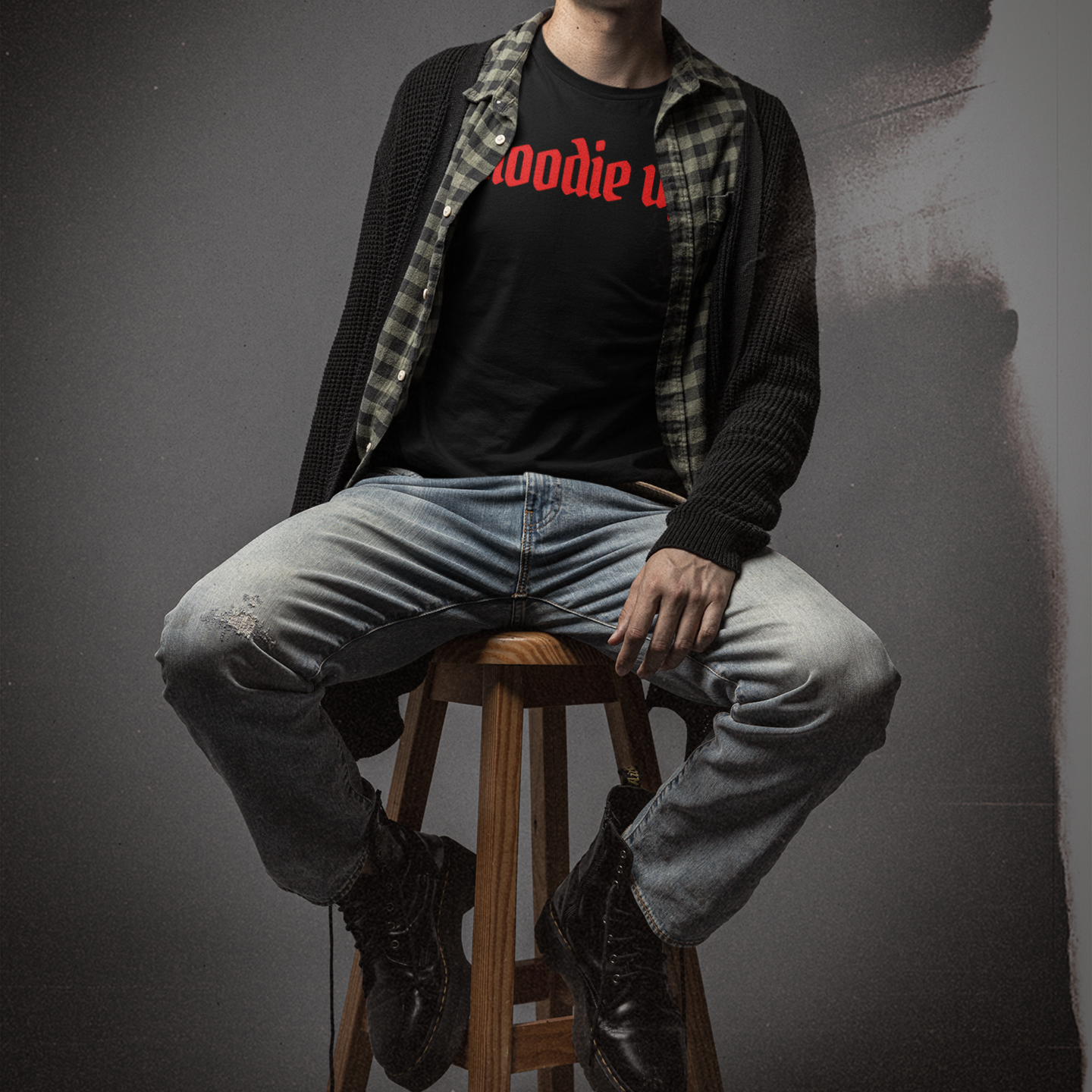 Person sitting on a stool wearing a black shirt with red text, gray pants, and a plaid jacket.