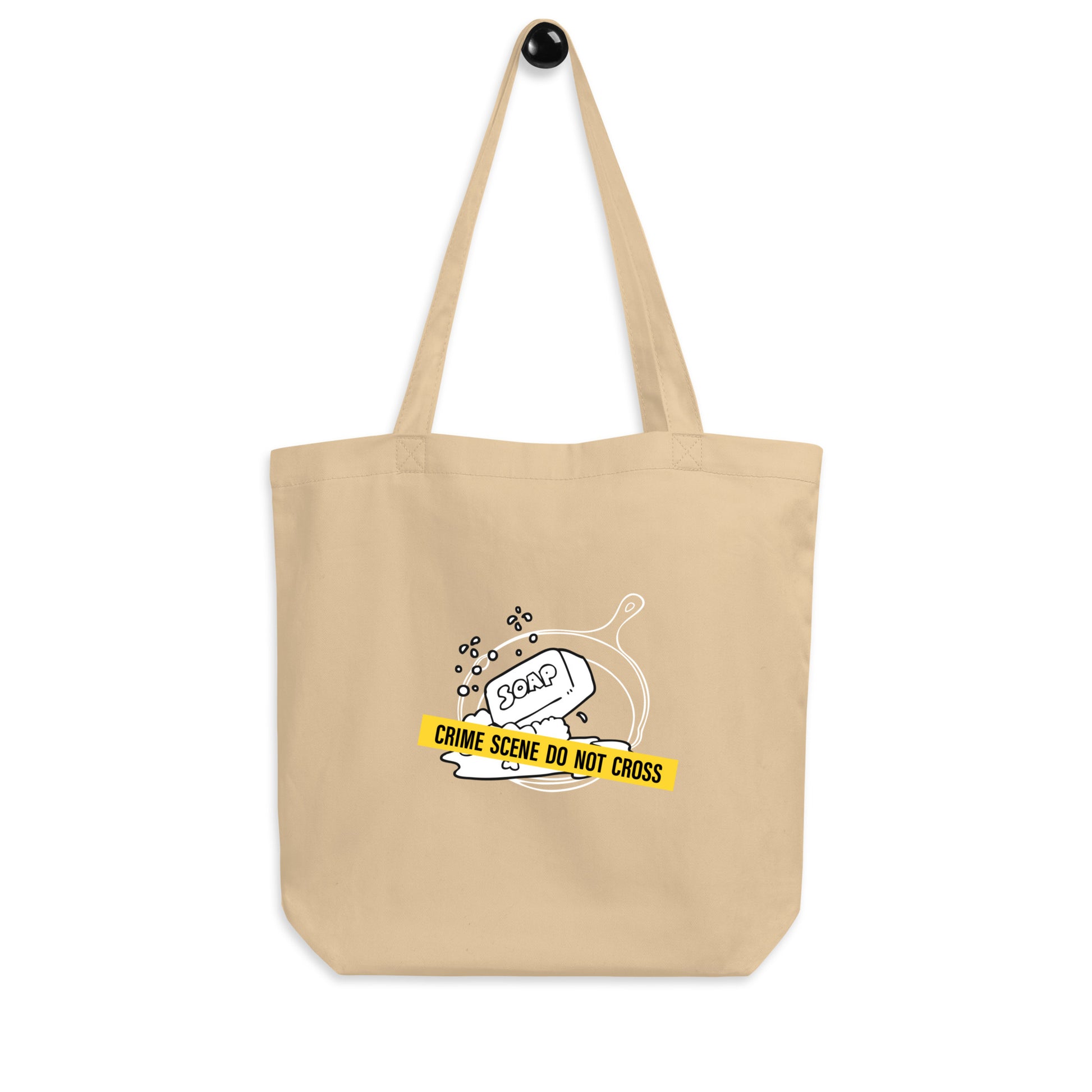 An oyster beige colored cotton tote bag featuring a graphic of a cast iron skillet filled with soap suds, blocked by a yellow strip of tape reading 'CRIME SCENE DO NOT CROSS'.