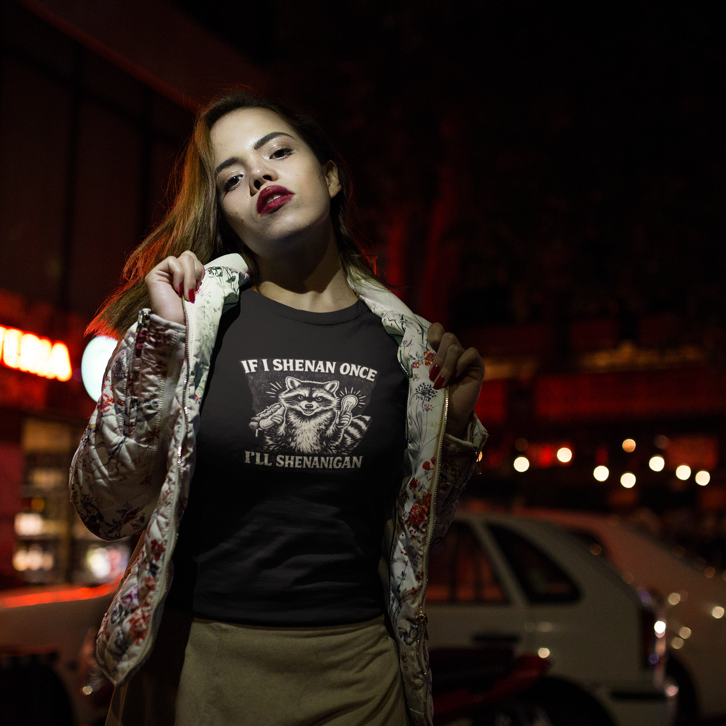A woman wearing a red, blue and green floral print jacket with her hands at both collars holding the jacket open to reveal her graphit colored short-sleeve crewneck t-shirt featuring a distressed black and white illustration of a smiling raccoon holding a hot dog in one paw and a glowing lightbulb in the other. Arched above the raccoon is text that reads 'IF I SHENAN ONCE', and below the raccoon is text flanked by stars that reads 'I'LL SHENANIGAN'. It is night and she is standing in front of parked cars.