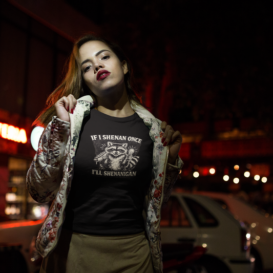 A woman wearing a red, blue and green floral print jacket with her hands at both collars holding the jacket open to reveal her graphit colored short-sleeve crewneck t-shirt featuring a distressed black and white illustration of a smiling raccoon holding a hot dog in one paw and a glowing lightbulb in the other. Arched above the raccoon is text that reads 'IF I SHENAN ONCE', and below the raccoon is text flanked by stars that reads 'I'LL SHENANIGAN'. It is night and she is standing in front of parked cars.