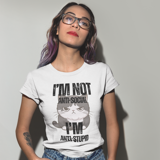 A woman wearing glasses and dyed purple hair tips, standing face the viewer with her head tilted to the left. She is wearing a white crew neck t-shirt featuring a graphic of a plump, annoyed looking cat and black, bold distressed all-caps text that reads 'I'M NOT ANTI-SOCIAL I'M ANTI STUPID'. 