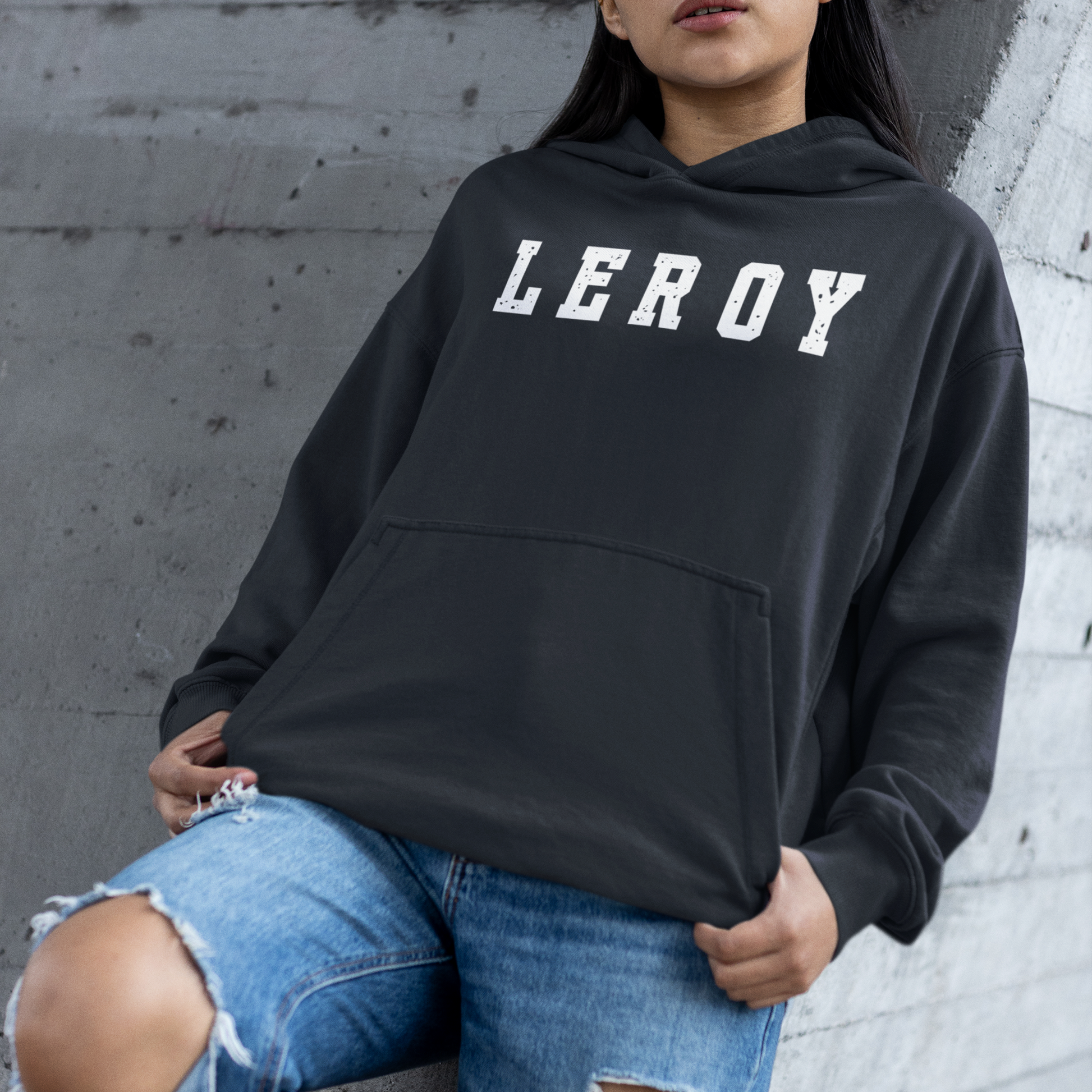 A woman wearing blue jeans torn at the knees leaning against a masonry wall. She is wearing a navy blue pullover hoodie featuring the name 'LEROY' printed horizontally across the chest in large, distressed white collegiate block letters. The hoodie includes a front kangaroo pocket and a ribbed waist and cuffs.