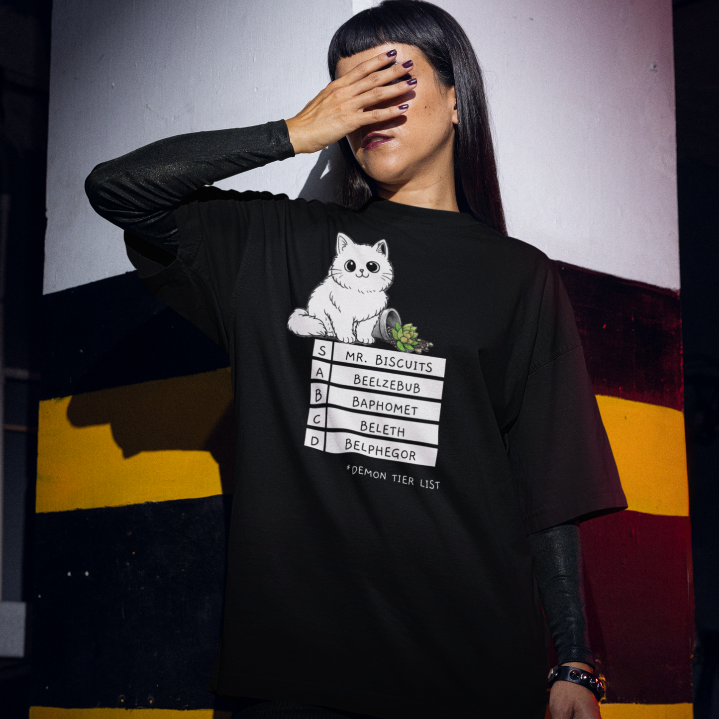 A Gothic styled woman with long, dark hair standing with her right hand covering her eyes wearing a black colored t-shirt with a graphic of a white, fluffy kitten standing behind a knocked over plant. Beneath the cat is a 'Demon Tier List' with the cat 'Mr. Biscuits' listed atop the list in the S tier position.