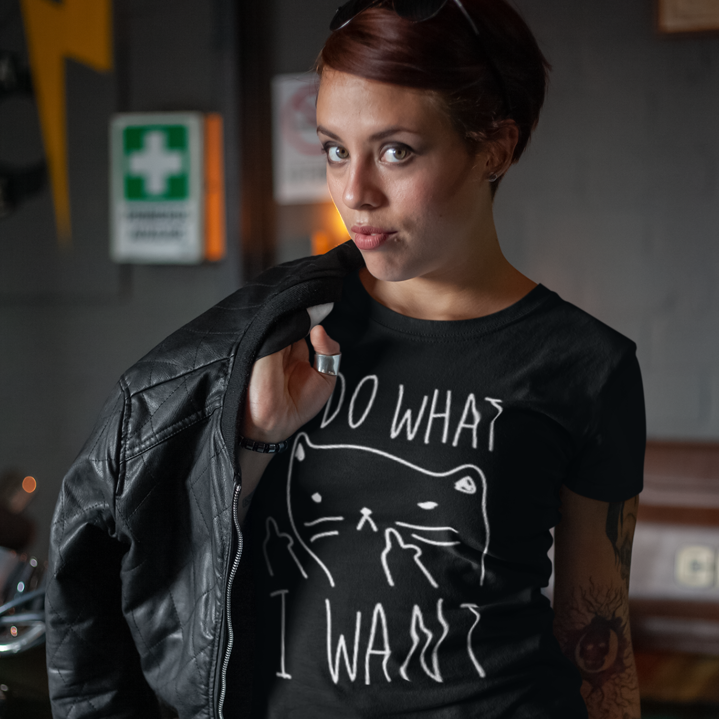 A woman with a leather jacket slung over her right shoulder and a sleeve tattoo on her left arm looking at the viewer while wearing a black t-shirt with a graphic of a cat with anthropomorphic middle fingers raised to the viewer and text 'I DO WHAT I WANT' in an indoor setting with a First Aid kit visible in the background. 