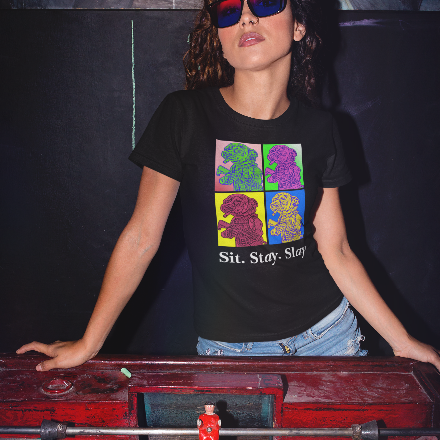 A woman wearing red and blue lensed sunglasses leaning on the edge of a foosball table with only the her goal end in view. She has long dark curly hair and pouty lips. She is wearing s black colored women's t-shirt with a four-panel pop-art graphic of the tactical Chihuahua meme, with the text 'Sit. Stay. Slay' underneath as a tribute to the New Order album 'Technique' pop art style  cover art and jean shorts with only the waist visible above the table.