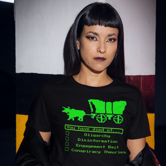 A goth-styled woman with jet black hair standing in front of a gray, maroon and gold painted wall. She is wearing A navy colored short-sleeve crewneck t-shirt featuring a bright green pixel art graphic referencing retro video games. The design depicts a covered wagon pulled by an ox. Below it, a text box in green pixel font reads 'You have died of...'. Below the text is a checklist with empty square boxes next to the options: 'Oligarchy', 'Disinformation', 'Engagement Bait', and 'Conspiracy Theories'.