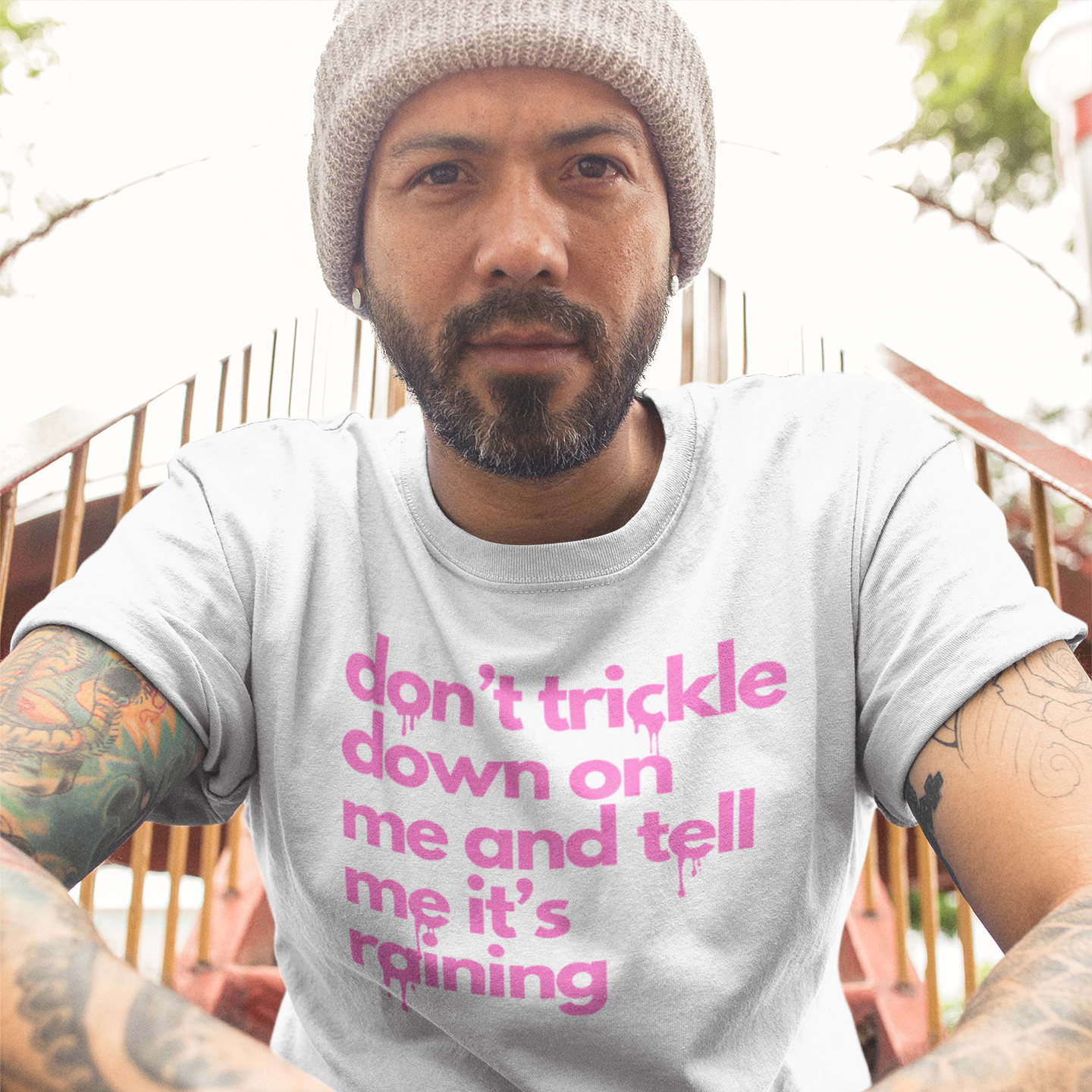 A bearded man sitting on a staircase leaning toward the viewer with his hands folded to the front. He has a full beard and is wearing a white colored t-shirt with pink text reading 'don't trickle down on me and tell me it's raining' in lower case letters with a dripping effect on some of the letters.