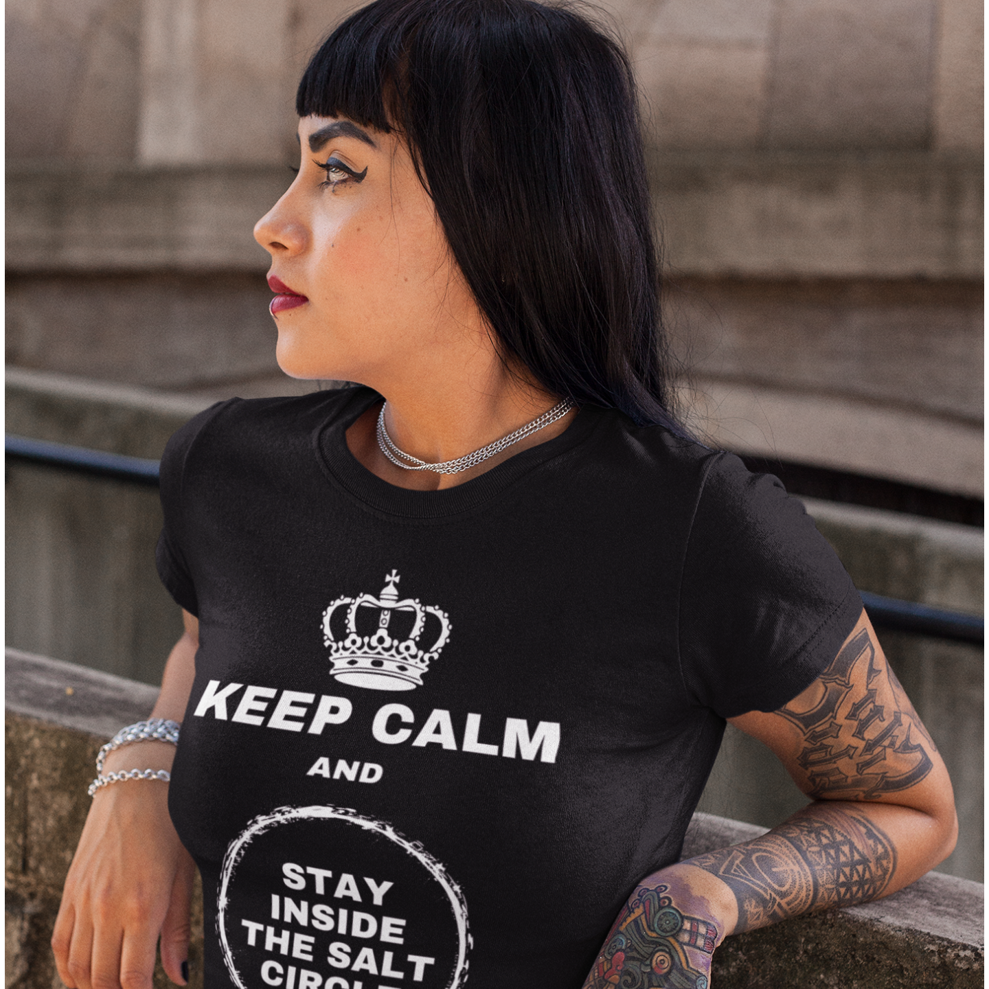 A dark haired, Gothic styled woman leaning against a wall with her head turned to her right and a tattoo covered left arm wearing a black colored t-shirt with a crown graphic over top of bold all-caps text that reads 'KEEP CALM AND STAY INSIDE THE SALT CIRCLE, with a white circular graphic to represent a 'salt circle'. 