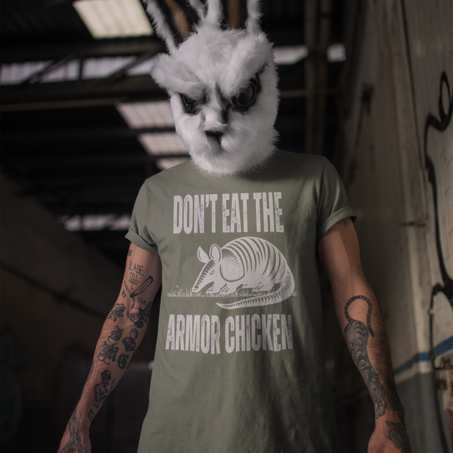 A man with heavily tattooed arm wearing a frightening rabbit head costume standing in a graffit covered hallway. He is wearing a moss green t-shirt with bold, all-caps, distressed white text that says 'DONT EAT THE ARMOR CHICKEN'. The text is separated into two lines by a white, line drawing graphic of an armadillo eating grass. 