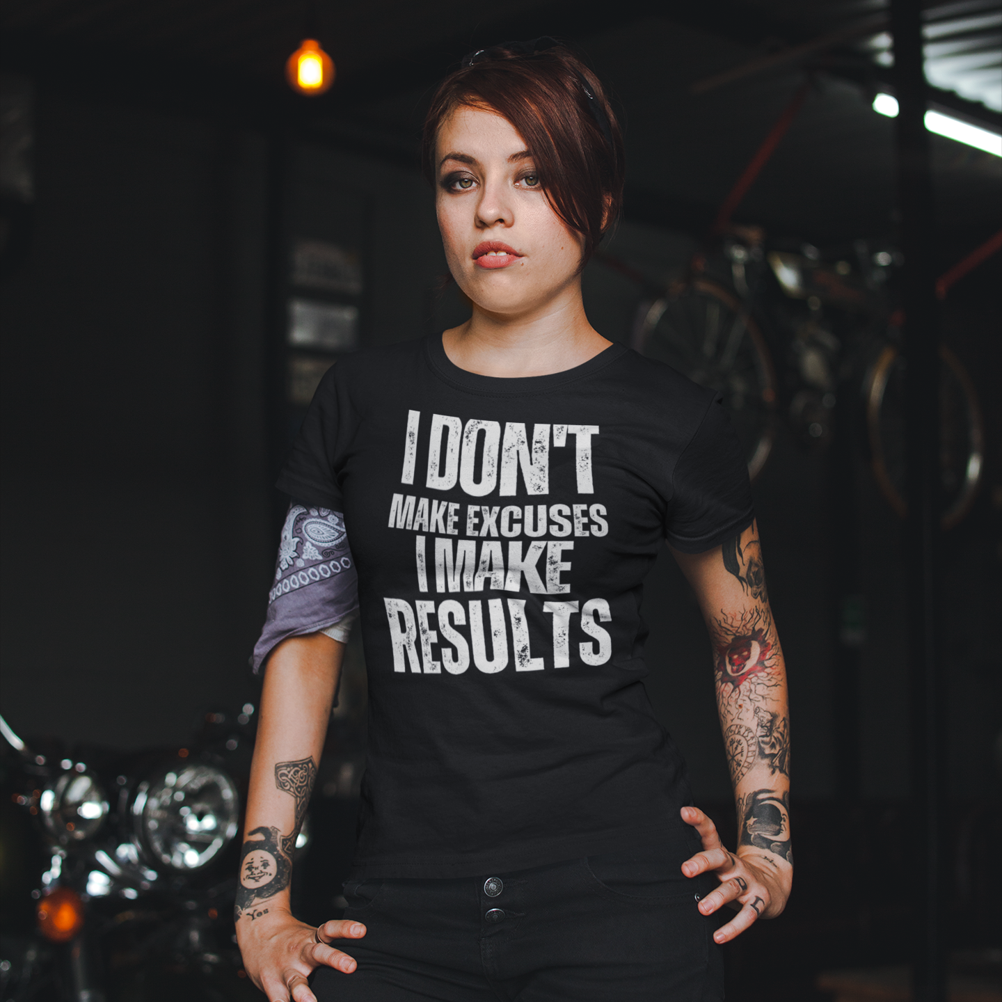 A woman with sleeve tattoos standing in front of a motorcycle wearing a black t-shirt with bold, all-caps, distressed text that reads 'I DON'T MAKE EXCUSES I MAKE RESULTS', on a white background.