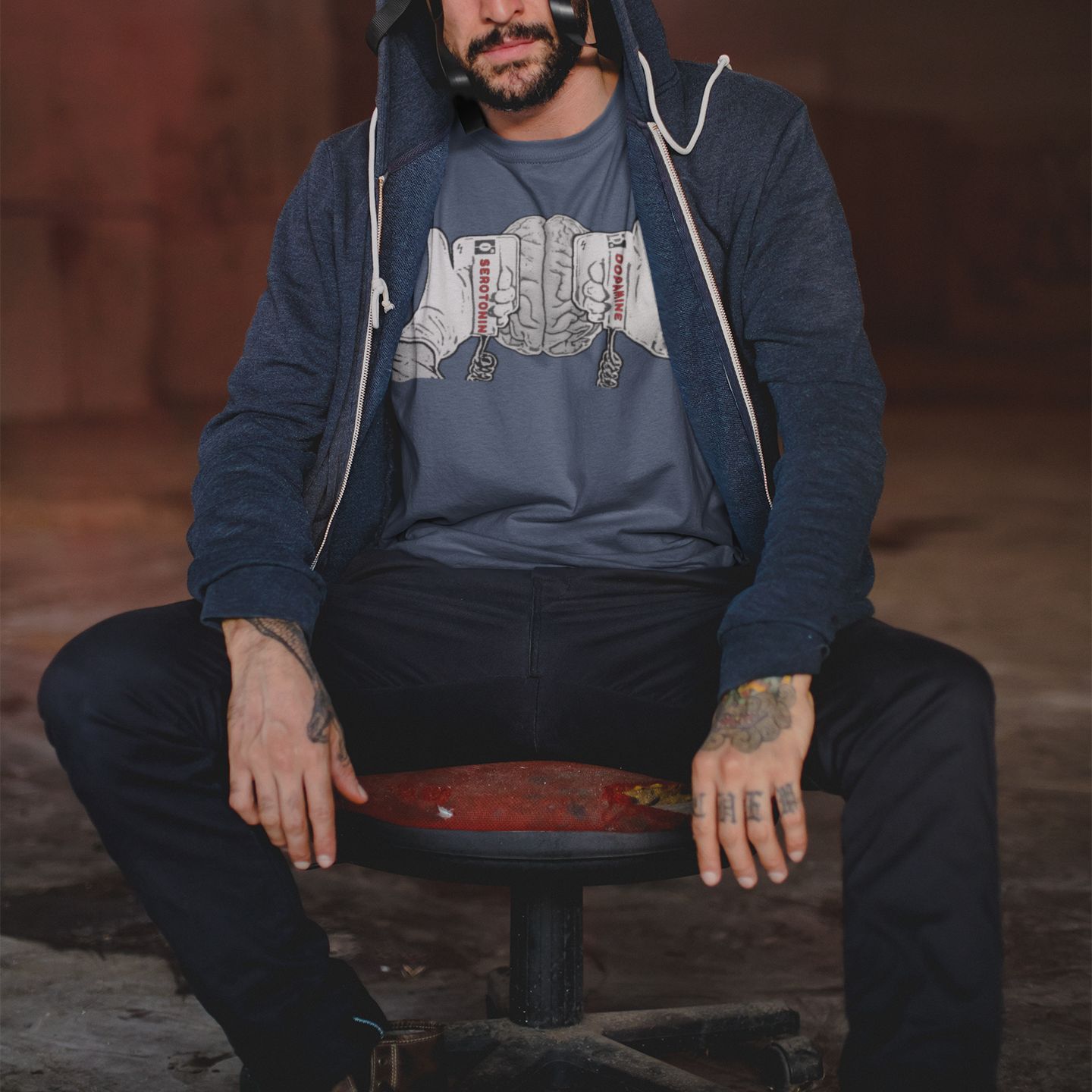 A man with tattooed hands sitting on a stool facing the viewer. He is wearing a blue t-shirt with a black and white line drawing graphic on the chest of a brain beneath two hands holding defibrillating paddles. The left paddle is labelled Serotonin and the right paddle is labelled Dopamine.  