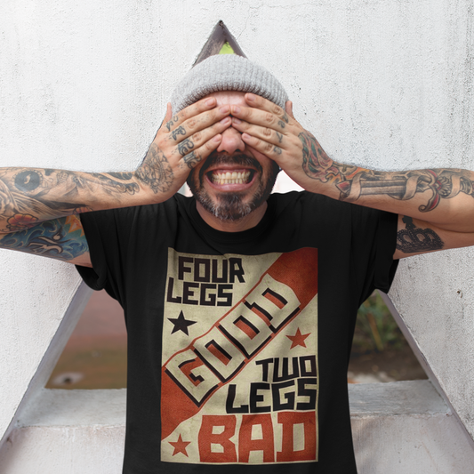 A man standing in front of a large, see through masonry triangle with his hands over his eyes. He is wearing a black colored short-sleeve crewneck t-shirt featuring a retro propaganda-style rectangular graphic. The design uses a textured tan, red, and black color palette with diagonal geometric blocking. It features the text 'FOUR LEGS' in black, a large diagonal 'GOOD' in off-white, 'TWO LEGS' in black, and a large 'BAD' in red at the bottom. The design is accented with small black and red stars.