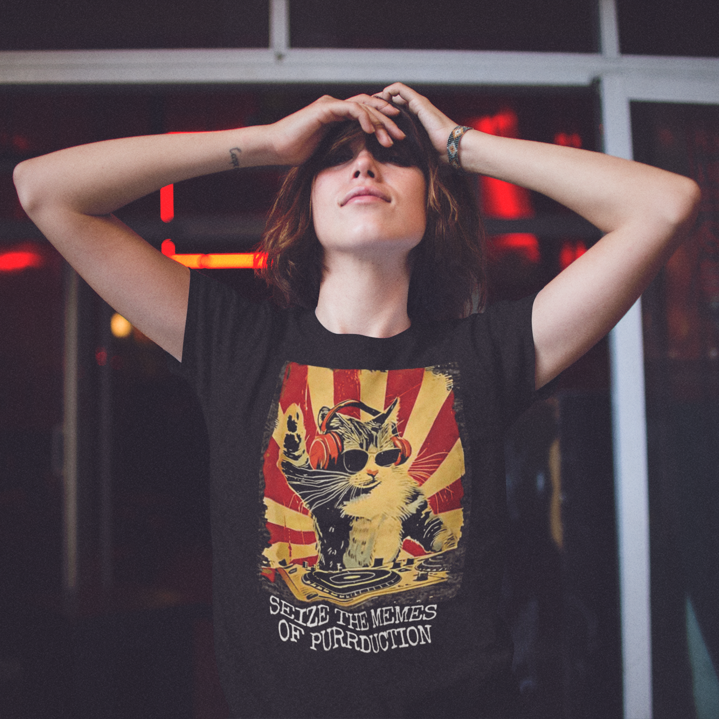 A woman standing against a darkened background with her arms raised and bent at the elbows, resting her hands against her forehead. Her head is tilted slight back and she has a mischevious grin. She is wearing a black t-shirt with a graphic of a cat DJing against a vintage propaganda style poster background, with the text "SEIZE THE MEMES OF PURRDUCTION" at the bottom