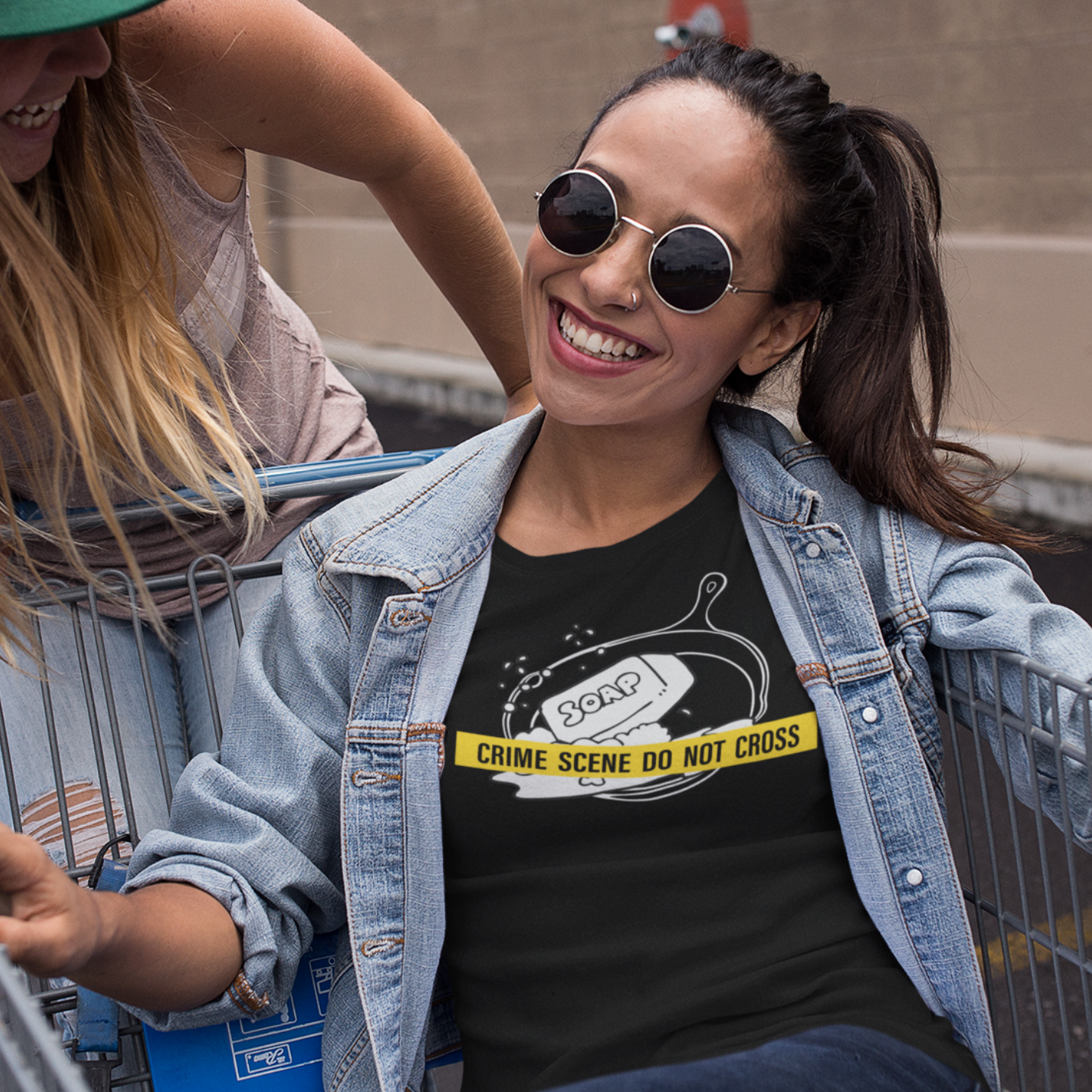 Two women in a shopping cart with one wearing sunglasses and a black t-shirt with a graphic of a cast iron pan with a bar of soap in it crossed with police style scene protection tape that reads - Crime Scene Do Not Cross.