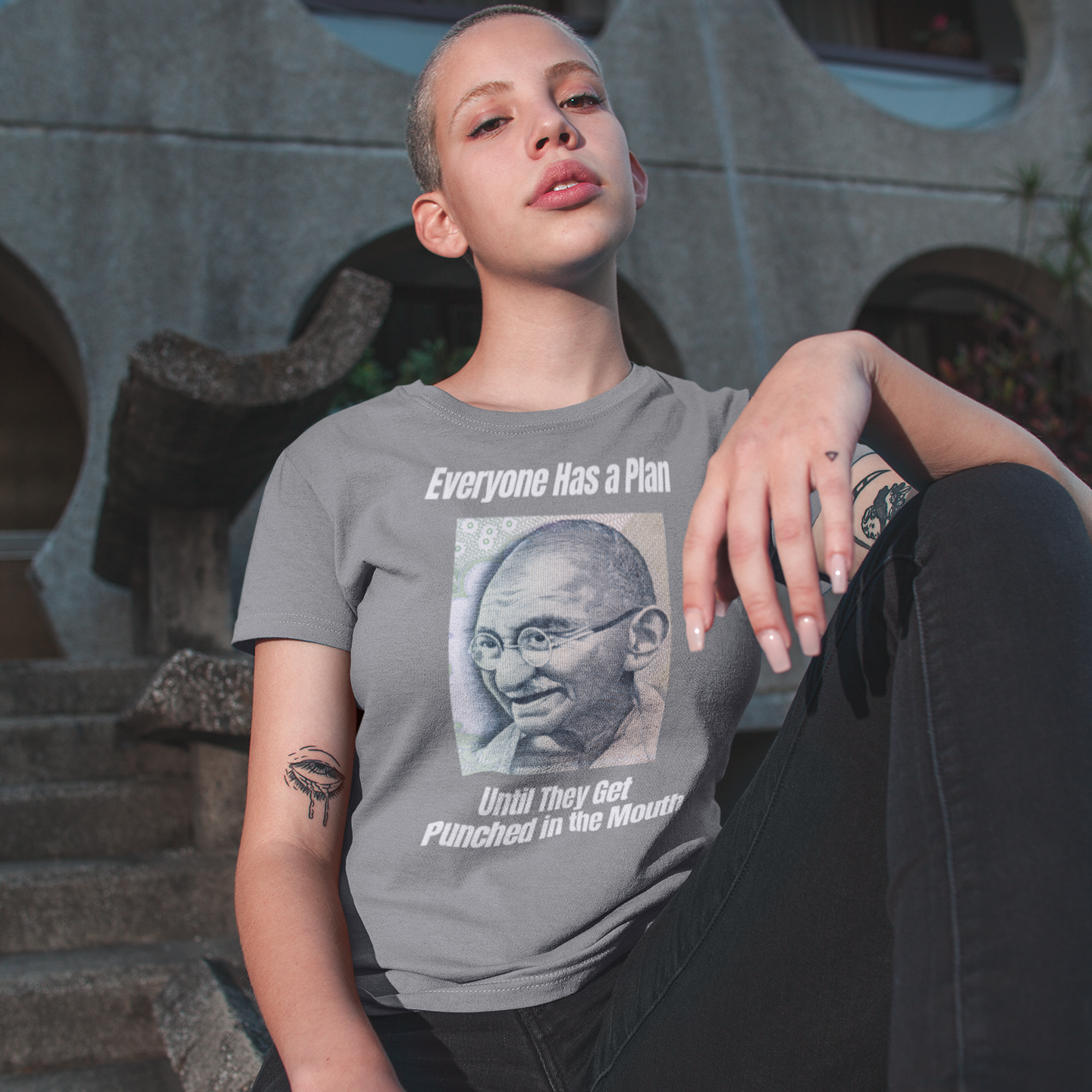 A woman with short cropped blonde hair sitting outdoors on concrete steps. Her left knee is raised in front of her chest and she is resting her left arm over her knee. She is wearing black jeans and a pepper colored tshirt featuring a vintage portrait of Mahatma Gandhi ironically paired with the famous quote 'Everyone Has a Plan Until They Get Punched in the Mouth'