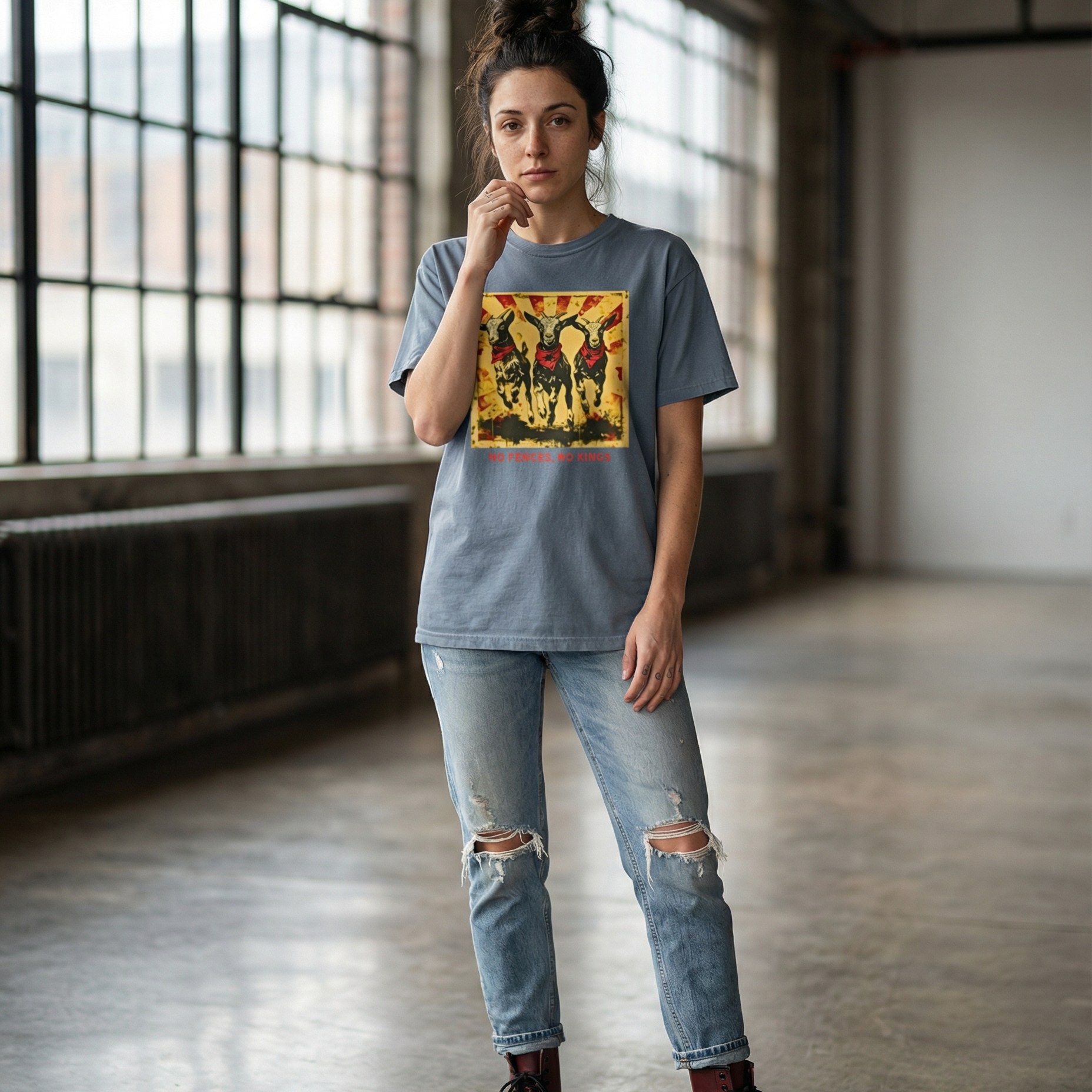 A woman standing in an empty industrial loft wearing distressed light-wash jeans and maroon Doc Martens. She is wearing a black colored Comfort Colors 1717 heavyweight t-shirt featuring a "No Fences, No Kings" graphic with three goats in red bandanas and a distressed sunburst background. Below the graphic is bold, red, all-caps text that reads, "NO FENCES, NO KINGS".