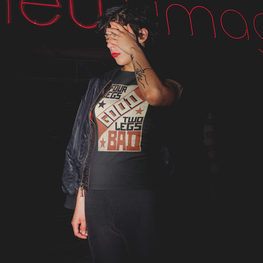 A woman standing against a black background with red neon lettering above her head. She is wearing a black colored short-sleeve crewneck t-shirt featuring a retro propaganda-style rectangular graphic. The design uses a textured tan, red, and black color palette with diagonal geometric blocking. It features the text 'FOUR LEGS' in black, a large diagonal 'GOOD' in off-white, 'TWO LEGS' in black, and a large 'BAD' in red at the bottom. The design is accented with small black and red stars.